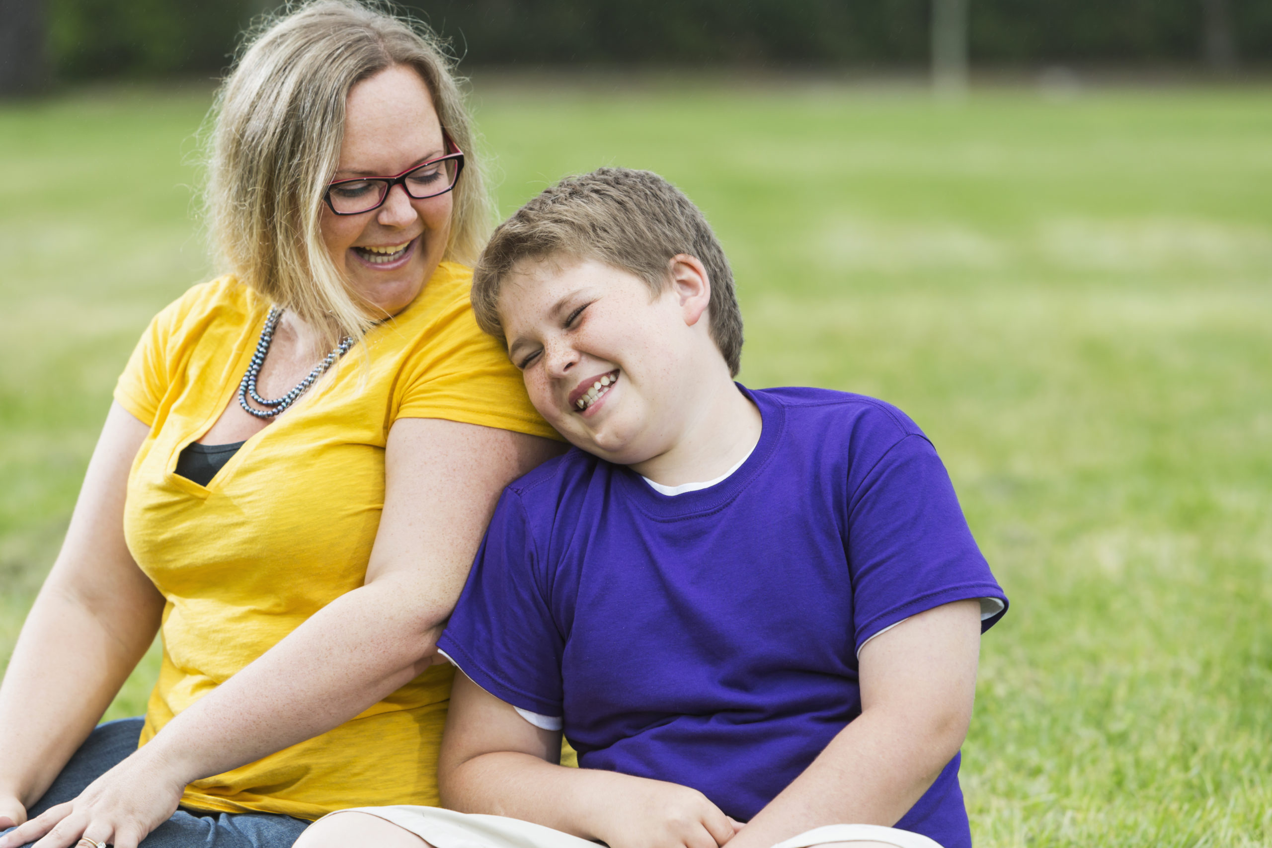 Mother and son laughing in the park Mother and preteen son sitting together on the grass in the park, laughing.