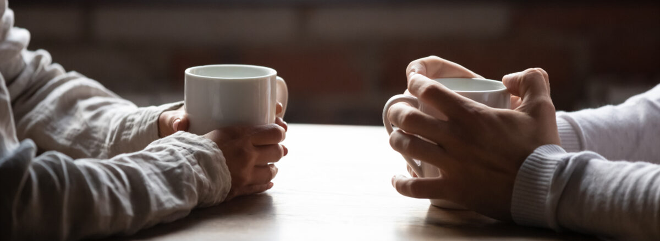 Two people sitting across from each other at a table, they are holding white cups People holding coffee cups